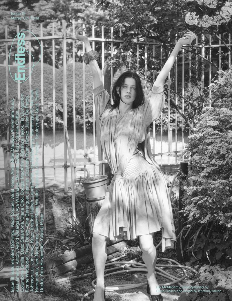 Fashion editorial shot of a young adult female model with arms raised, wearing a dress in an outdoor garden setting, black and white.