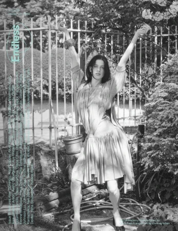 Fashion editorial shot of a young adult female model with arms raised, wearing a dress in an outdoor garden setting, black and white.