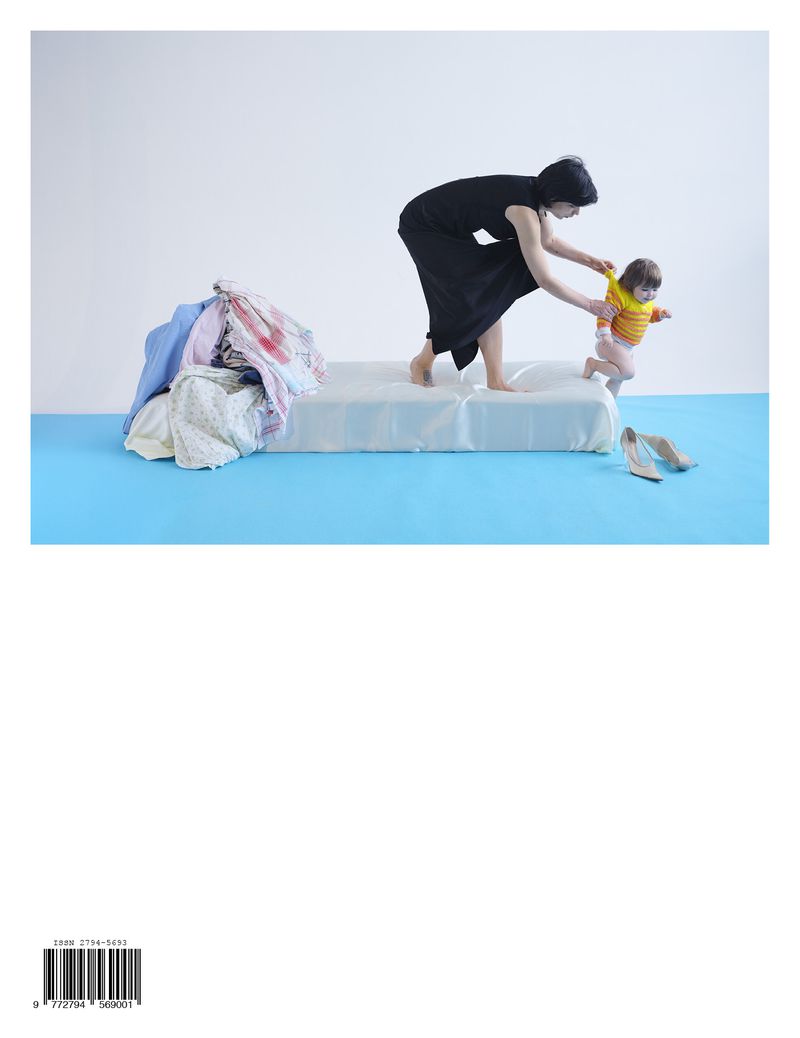Woman helps toddler walk on bed in minimalist studio. Laundry pile and high heels present, symbolizing motherhood, family, and home life.
