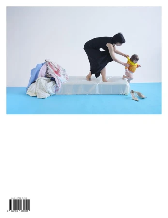 Woman helps toddler walk on bed in minimalist studio. Laundry pile and high heels present, symbolizing motherhood, family, and home life.