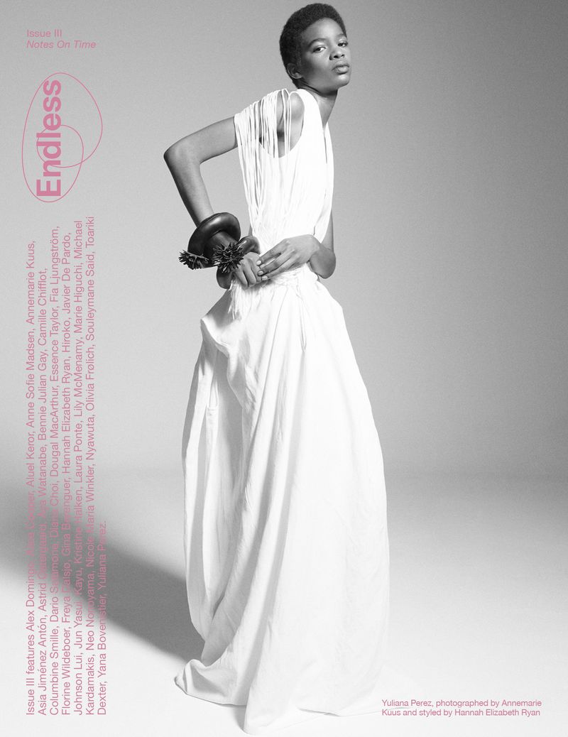 Black and white fashion shot of a Black model in a flowing white dress with a striking bracelet, set against a minimalist grey backdrop.