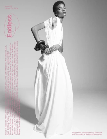 Black and white fashion shot of a Black model in a flowing white dress with a striking bracelet, set against a minimalist grey backdrop.