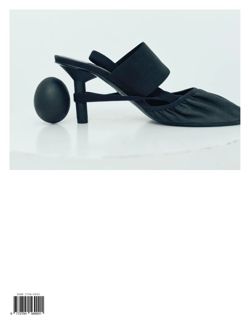 A black pointed low-heel mule shoe balancing beside a matte black egg on a plain white surface.