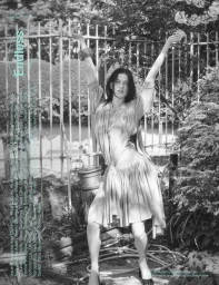 Black and white editorial fashion image of a model with arms raised in a lush garden setting with an iron fence background.