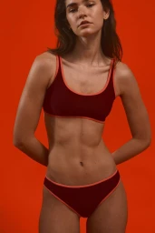 Female model posing in a burgundy two-piece bikini with contrasting orange trim against a solid orange background.