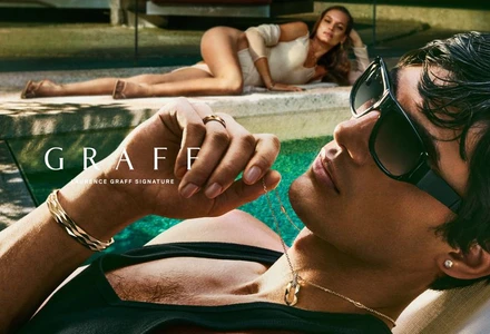 Model Akbar Shamji wearing black sunglasses and gold Graff jewelry poolside with Faretta reclining in the background.