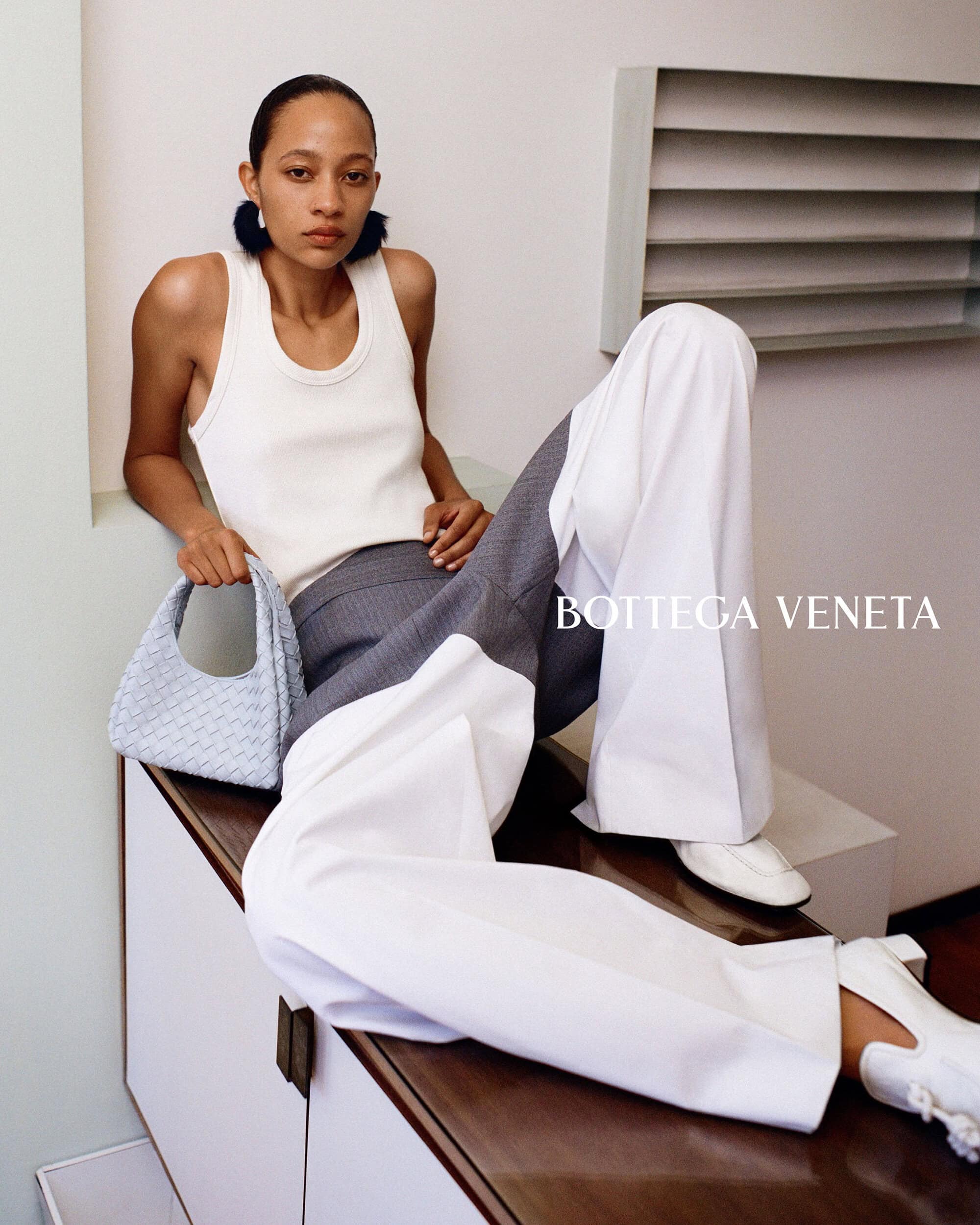Model wearing a white tank top and wide trousers holding a light blue woven handbag while seated on a counter