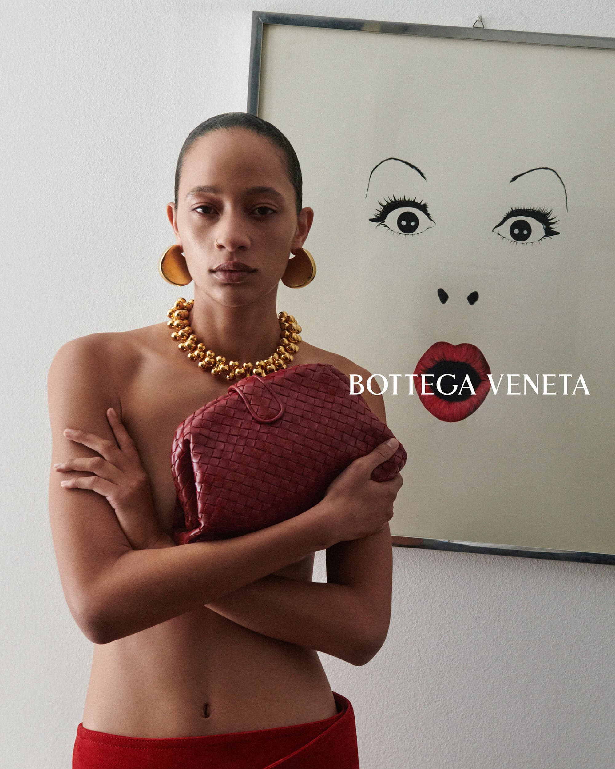 Selena Forrest holding a burgundy woven clutch and wearing chunky gold jewelry beside framed artwork for Bottega Veneta.