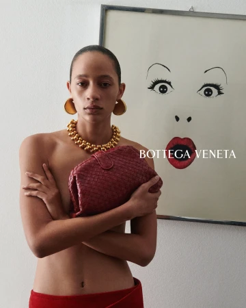 Selena Forrest holding a burgundy woven clutch and wearing chunky gold jewelry beside framed artwork for Bottega Veneta.