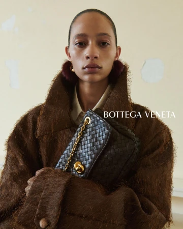Model Selena Forrest in a textured brown coat holding a black woven Bottega Veneta shoulder bag with gold hardware.