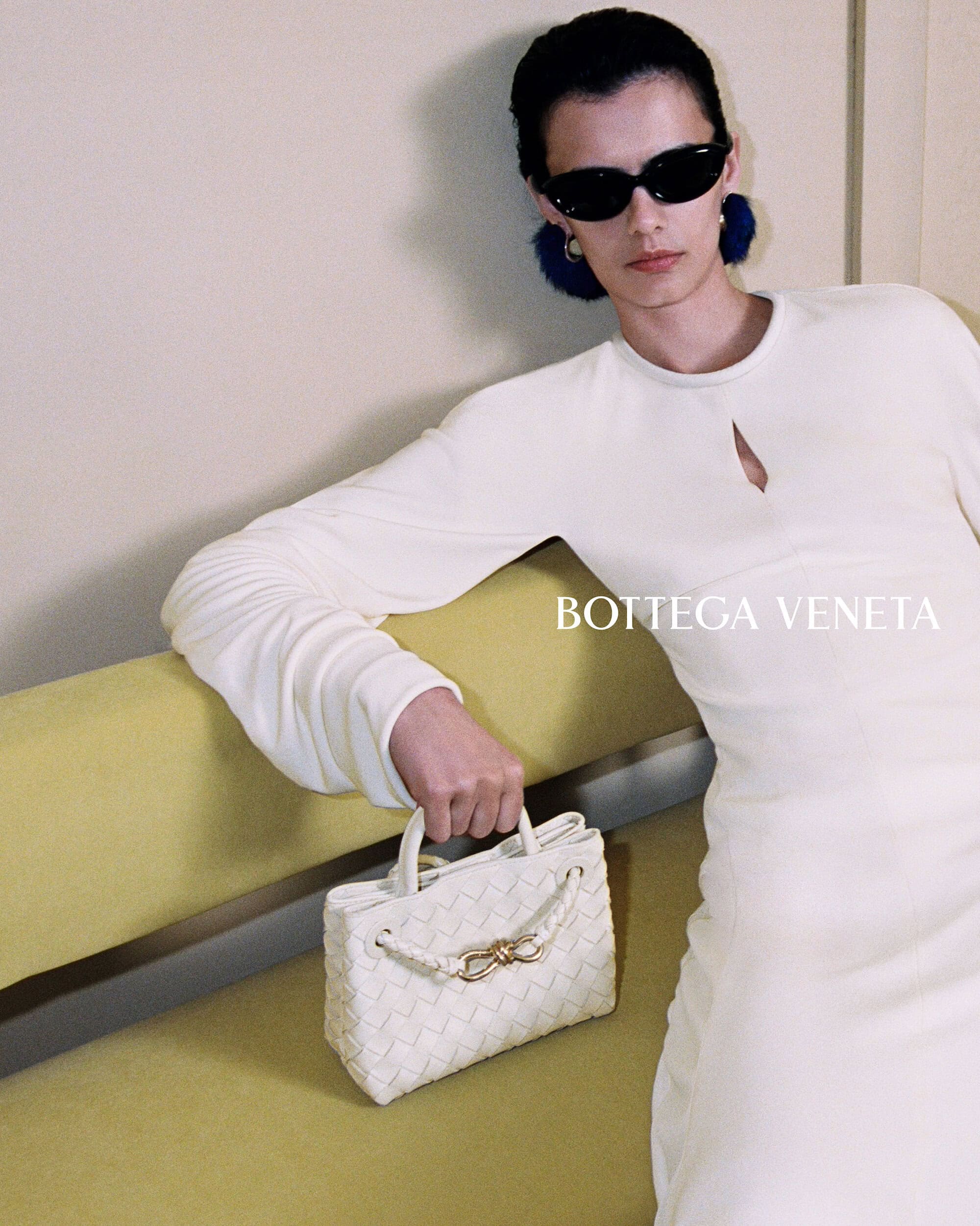 Model wearing a white dress and black sunglasses holding a white woven leather handbag while resting on a yellow sofa.