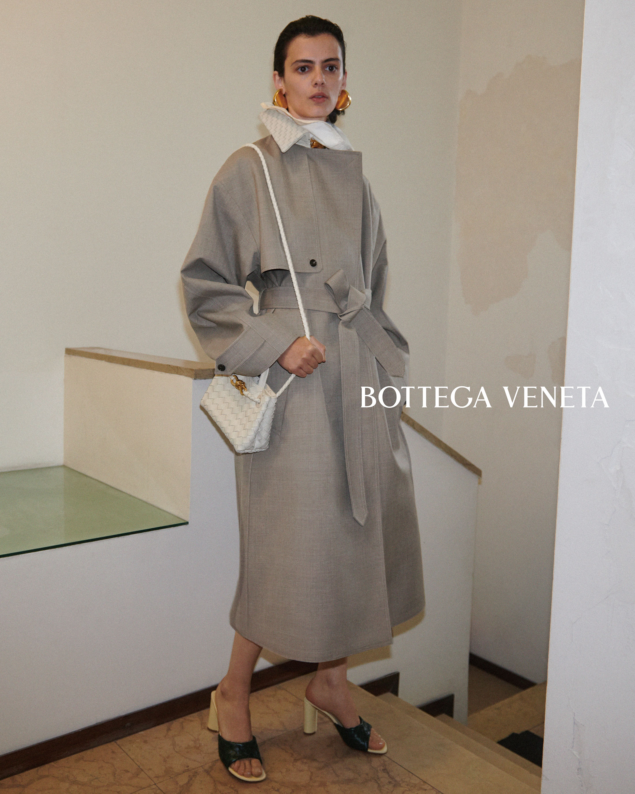 Model wearing a belted taupe trench coat and carrying a small white crossbody handbag on a staircase for Bottega Veneta.