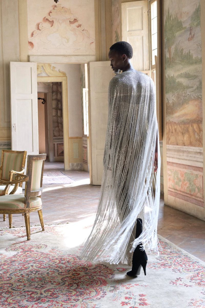 Model wearing a floor-length silver fringed beaded cape and black high-heeled boots in a historic room with frescoed walls.