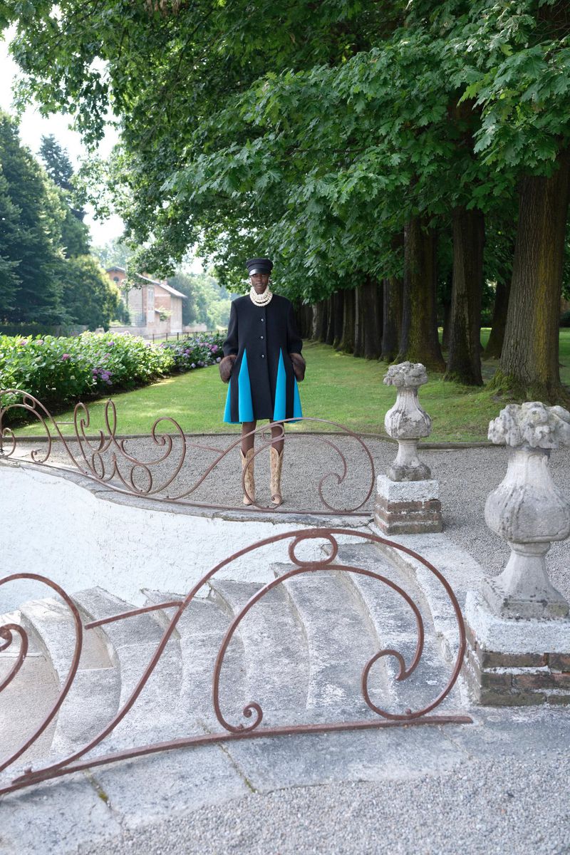 Model posing in a black flared coat with bright blue inserts atop ornate stone steps in a lush estate garden.