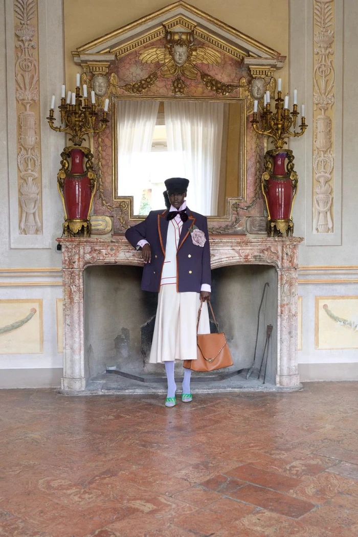 Model in a navy tailored blazer, white pleated skirt, and hat holding a tan leather bag in front of an ornate marble fireplace.