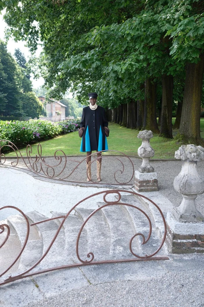 Model posing in a black flared coat with bright blue inserts atop ornate stone steps in a lush estate garden.