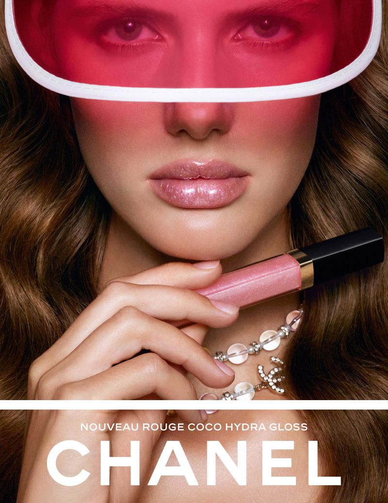 Model wearing a translucent pink visor and holding a tube of Chanel Rouge Coco Hydra Gloss to her lips.