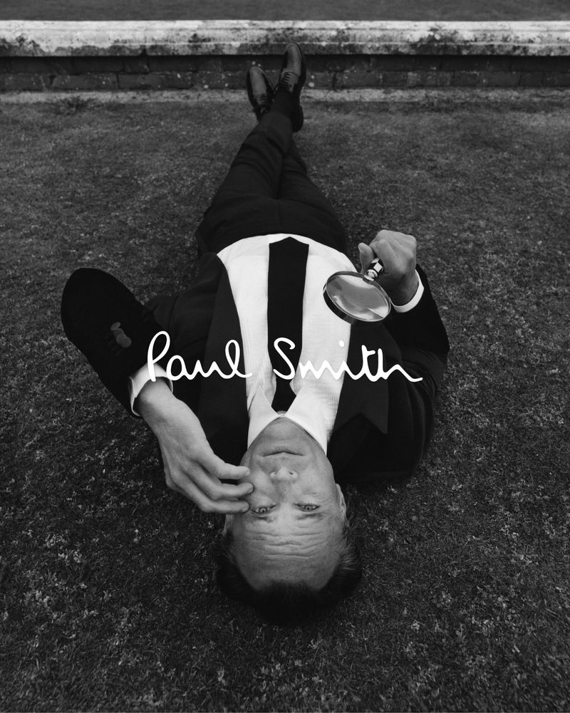Walton Goggins lying on grass in a black suit and white shirt holding a magnifying glass for Paul Smith.