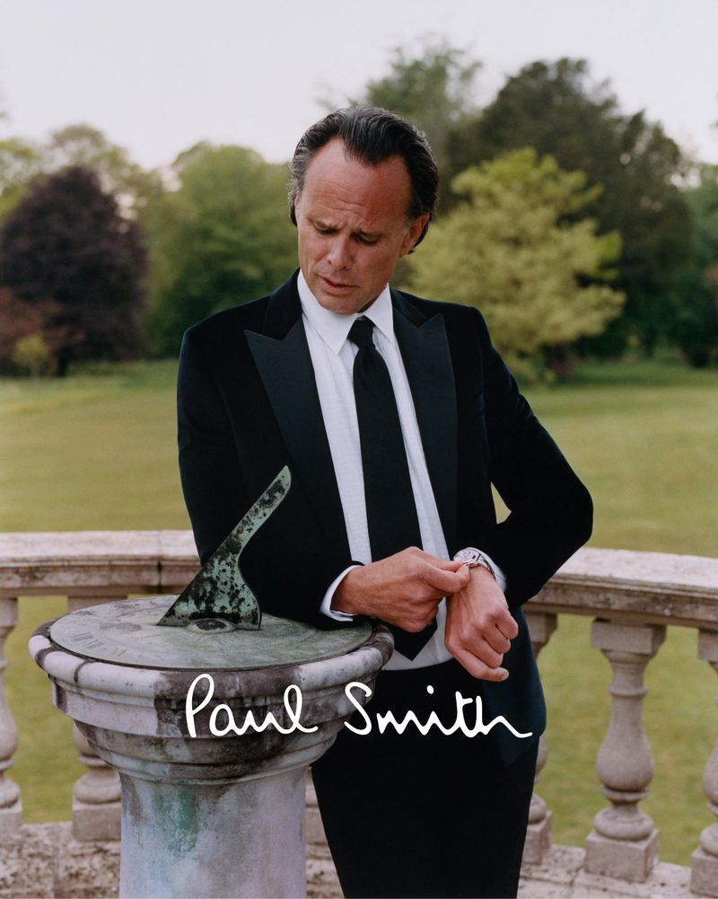 Walton Goggins wearing a black Paul Smith tailored suit while adjusting his watch next to a stone sundial outdoors.
