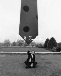 Walton Goggins seated on a lawn pointing up at a giant polka-dot tie in a black-and-white Paul Smith campaign photograph.