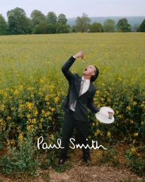 Walton Goggins standing in a yellow flower field wearing a tailored grey suit while eating a bite of cake from a fork.