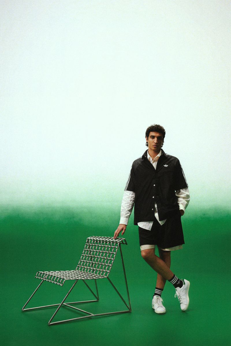 Model posing beside a wire mesh accent chair in an Adidas jacket, shorts, and Stan Smith sneakers against a green gradient backdrop.