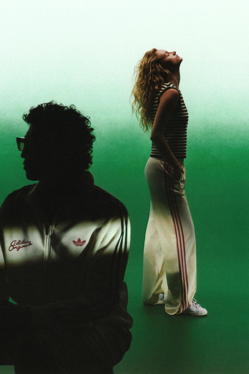 A woman in a striped top and cream track pants looking up beside a silhouetted man in an Adidas track jacket on a green backdrop.