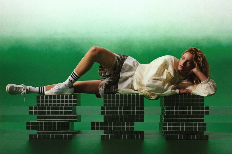Model reclining across green tiled plinths wearing an off-white jacket and white sneakers against a green gradient backdrop.