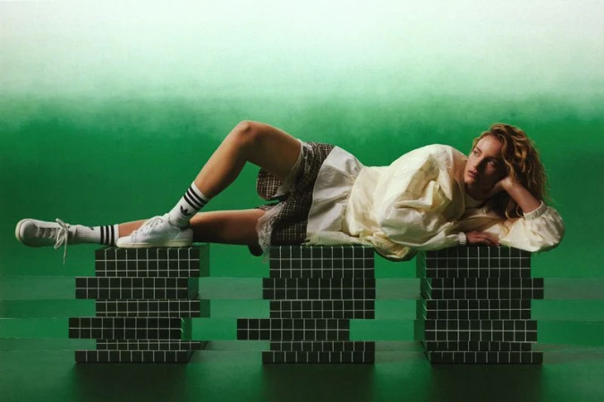 Model reclining across green tiled plinths wearing an off-white jacket and white sneakers against a green gradient backdrop.
