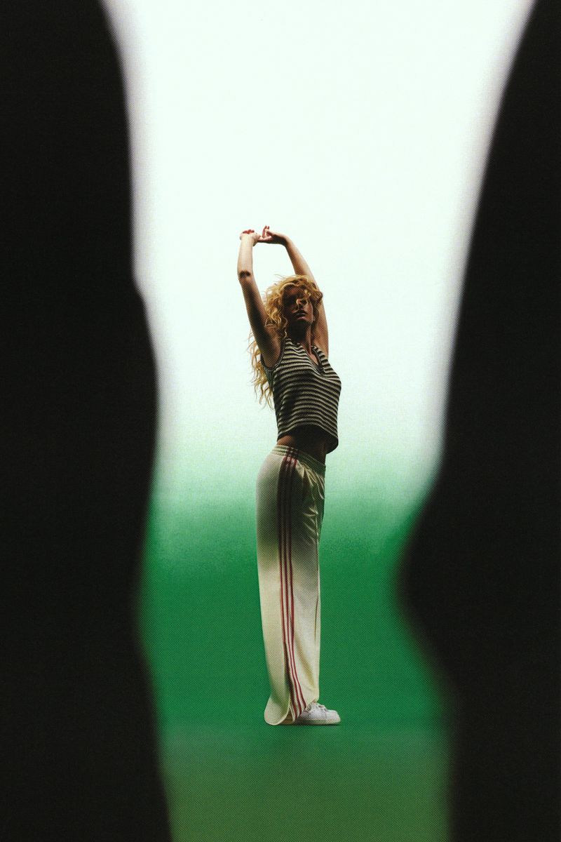 Model stretching with raised arms wearing striped top and cream track pants against a green gradient, framed by dark silhouettes.