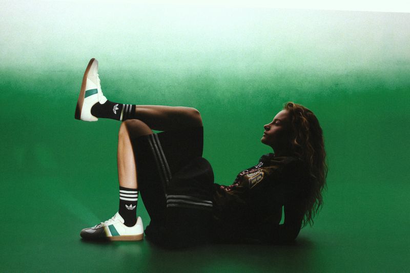 A woman reclines against a green gradient backdrop wearing black athletic shorts, black logo crew socks, and white low-top sneakers.