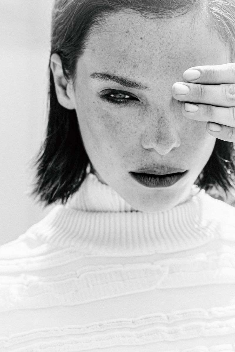 Black and white portrait of a freckled woman in a white turtleneck sweater covering one eye with her hand.