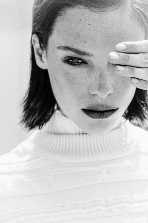 Black and white portrait of a freckled woman in a white turtleneck sweater covering one eye with her hand.