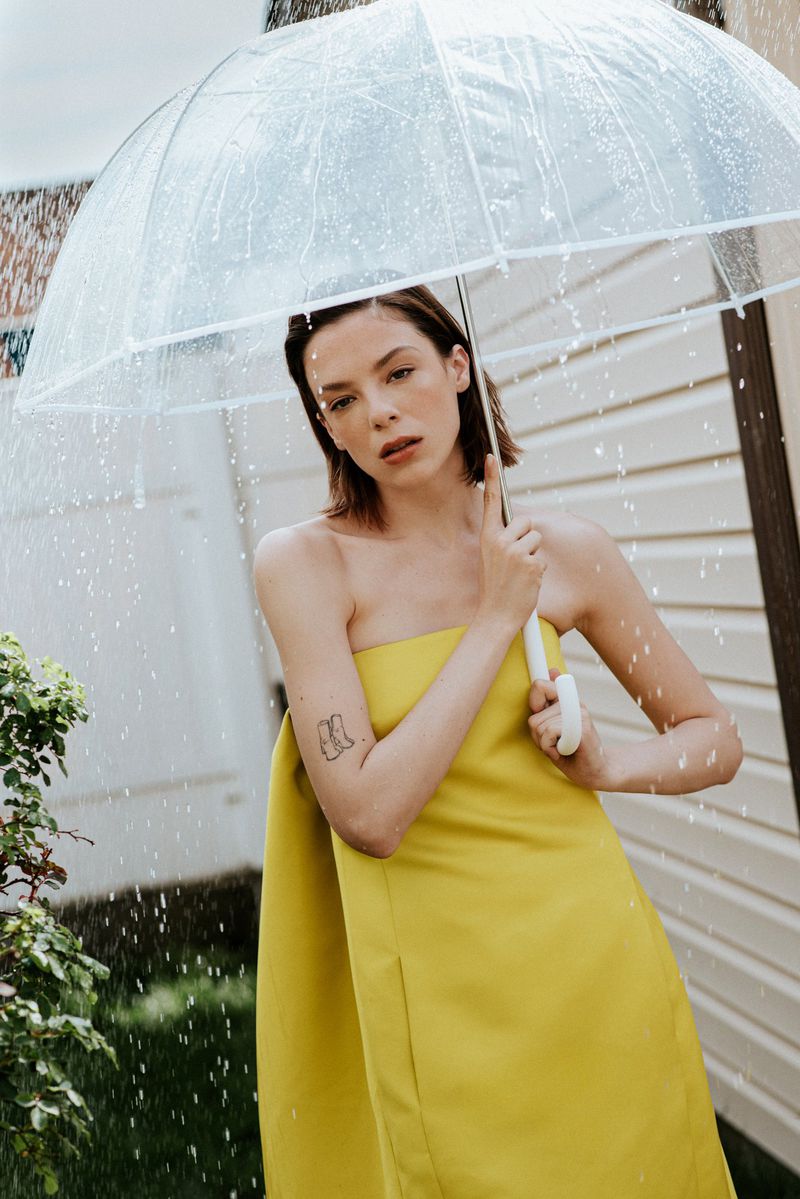 A woman in a strapless yellow dress holds a clear umbrella in the rain against a light-colored house exterior.