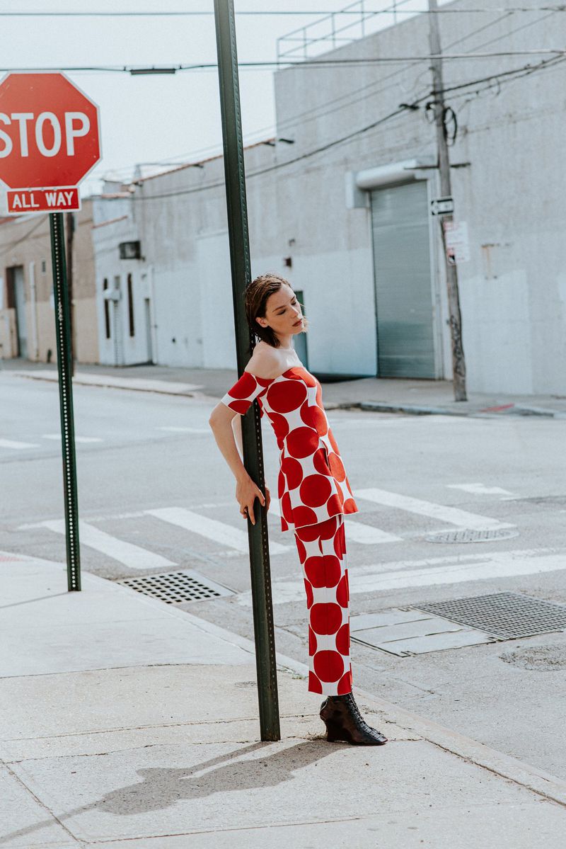 Woman in red and white polka dot outfit leaning against a street sign post on an urban street.