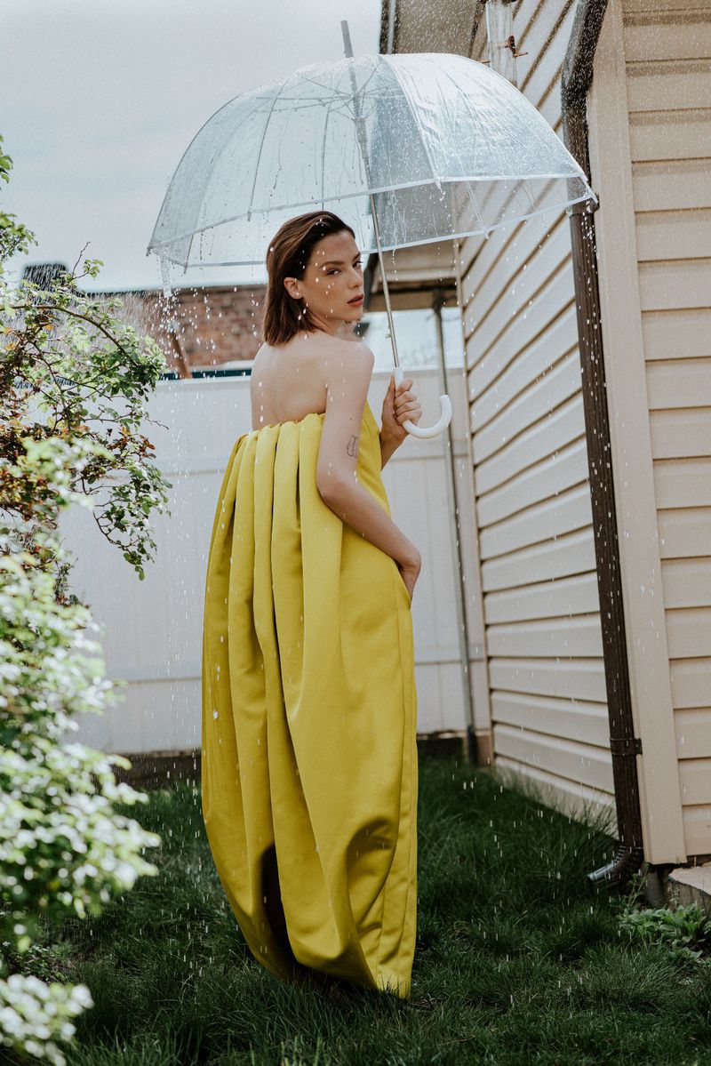 Woman in yellow dress holding a clear umbrella outdoors in the rain next to a house siding wall.