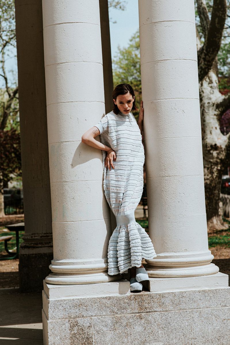 Woman in a textured light blue dress leaning between classical stone columns in an outdoor park setting.