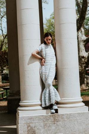 Woman in a textured light blue dress leaning between classical stone columns in an outdoor park setting.