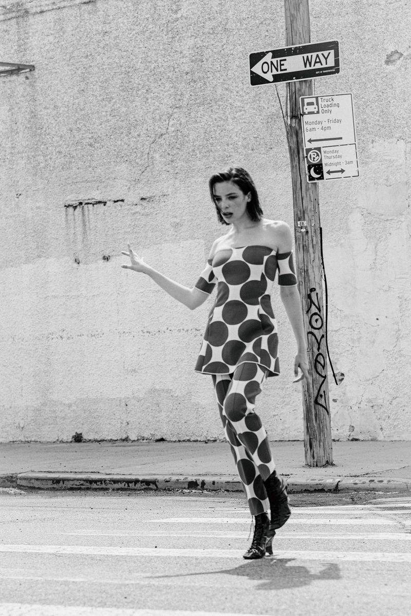 A model in a bold polka dot matching top and pants walks across a sunlit street next to a wooden utility pole in black and white.