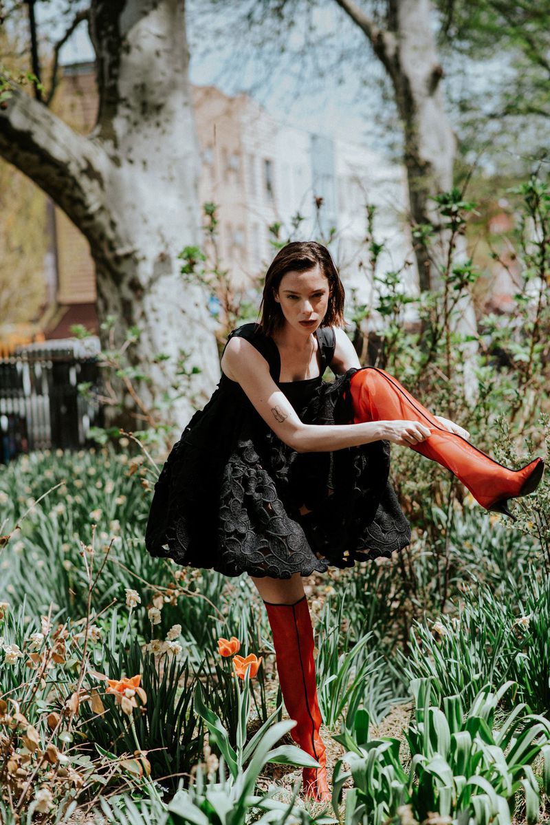 Woman in black dress and red boots adjusting footwear among trees and flowers for Tidal Magazine