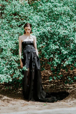Model wearing a strapless black gown with circular appliqués standing in front of lush green foliage in bright daylight.