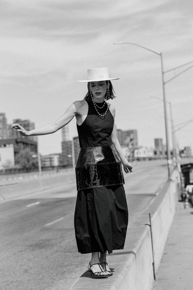 Model balancing on a concrete road barrier wearing a sleeveless black dress, glossy waist corset, white wide-brim hat, and sandals.