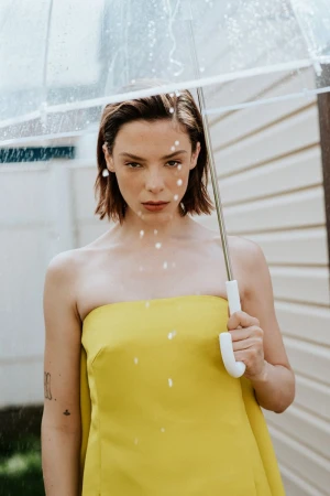 A woman in a strapless yellow dress holds a clear umbrella while rain falls outdoors against a siding wall.