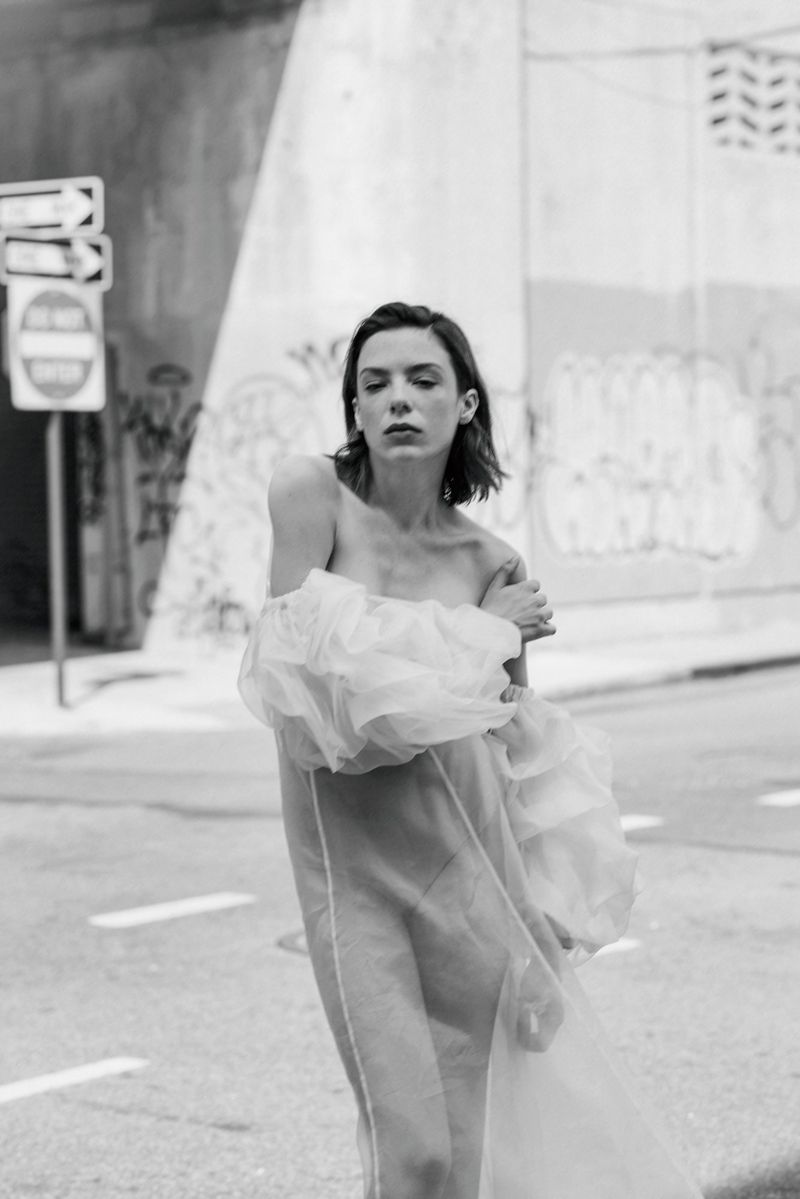 A woman wearing a sheer off-the-shoulder ruffled dress posing in black and white on an urban street.