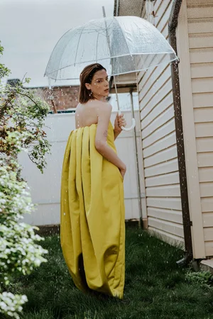 Woman in yellow dress holding a clear umbrella outdoors in the rain next to a house siding wall.