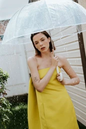A woman in a strapless yellow dress holds a clear umbrella in the rain against a light-colored house exterior.