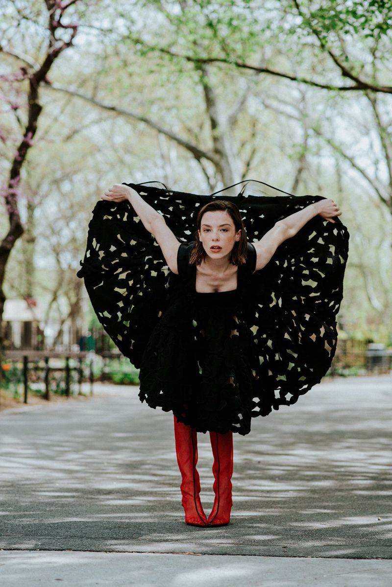 Woman in a voluminous perforated black dress and knee-high red boots posing on a paved park pathway.