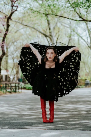 Woman in a voluminous perforated black dress and knee-high red boots posing on a paved park pathway.