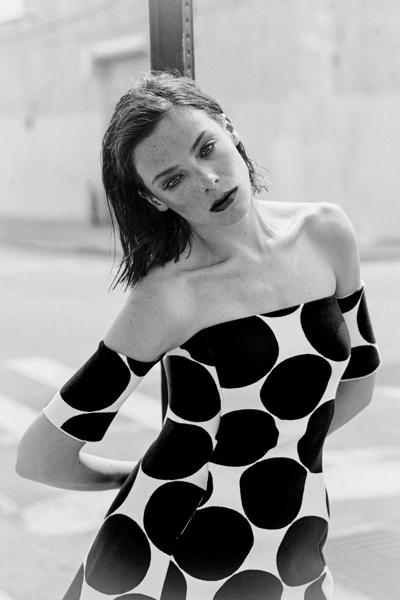Black and white portrait of a woman leaning against a pole wearing a bold off-the-shoulder polka dot dress outdoors.