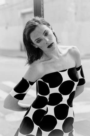 Black and white portrait of a woman leaning against a pole wearing a bold off-the-shoulder polka dot dress outdoors.